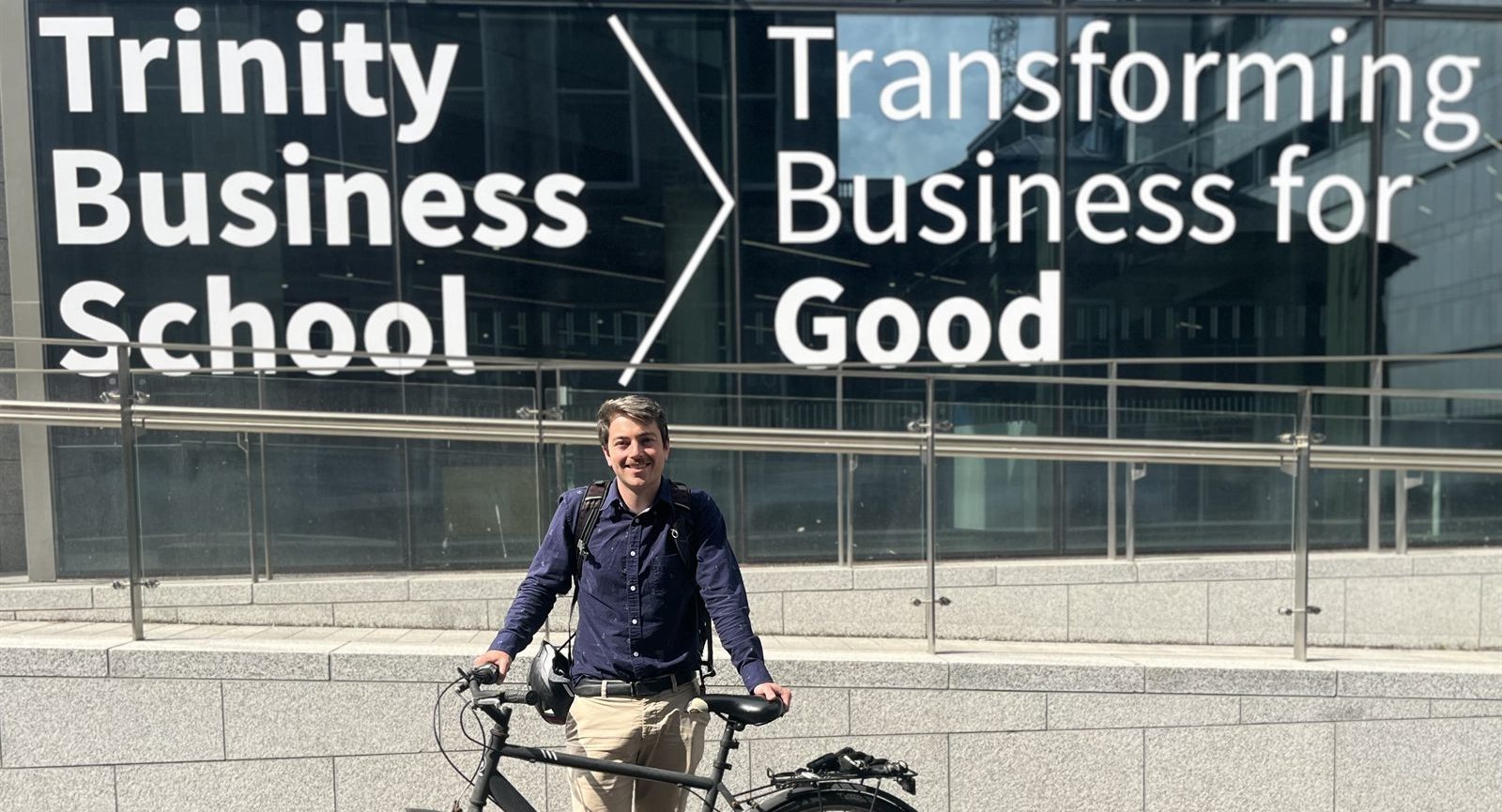 ElectrifyNow's founder standing with his commuter bike outside Trinity Business School, whose window reads "Transforming Business for Good."